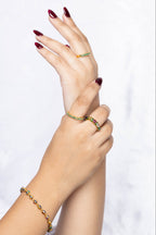A close-up of a person's hands and wrists wearing a gold bracelet and two matching gold rings with green gemstone accents, set against a soft white marble background.