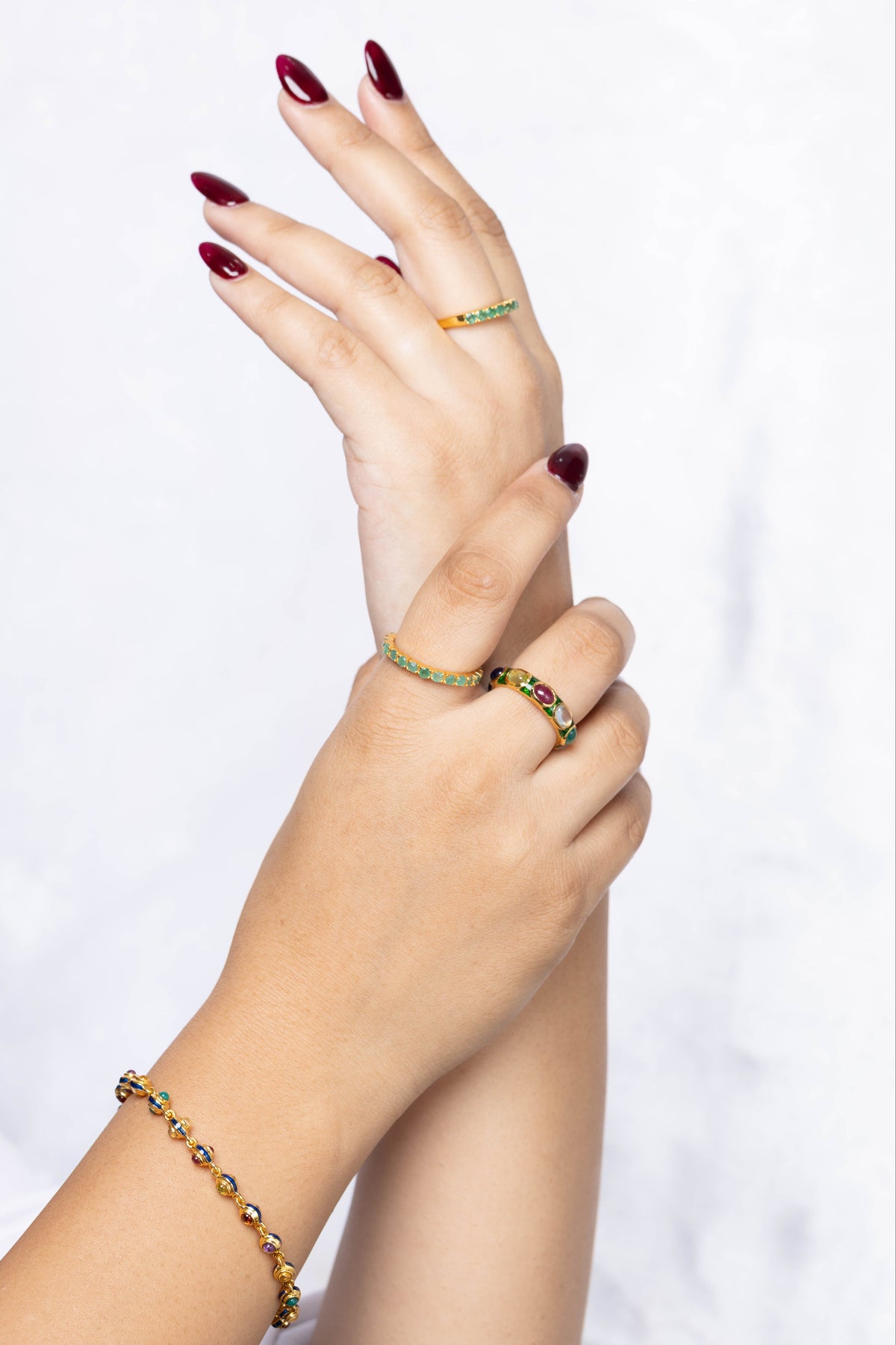 A close-up of a person's hands and wrists wearing a gold bracelet and two matching gold rings with green gemstone accents, set against a soft white marble background.