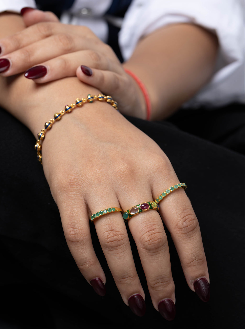 A close-up of a person's hands wearing gold rings and a matching gold bracelet, decorated with green and red gemstones. The person is wearing a white collared shirt and black trousers.