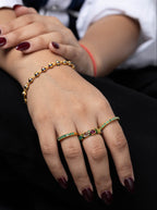 A close-up of a person's hands wearing gold rings and a matching gold bracelet, decorated with green and red gemstones. The person is wearing a white collared shirt and black trousers.