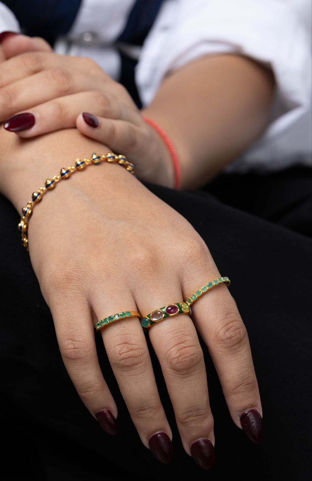 A close-up of a person's hands wearing gold rings and a matching gold bracelet, decorated with green and red gemstones. 
