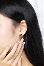A close-up of a woman with long black hair wearing a small gold stud earring with colorful gemstone accents, touching her cheek with her hand.