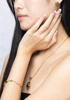A woman with long black hair models a matching gold jewelry set, including a delicate necklace with a dark amber and green stone pendant, a gold bracelet, and a small gold stud earring, against a light gray background.