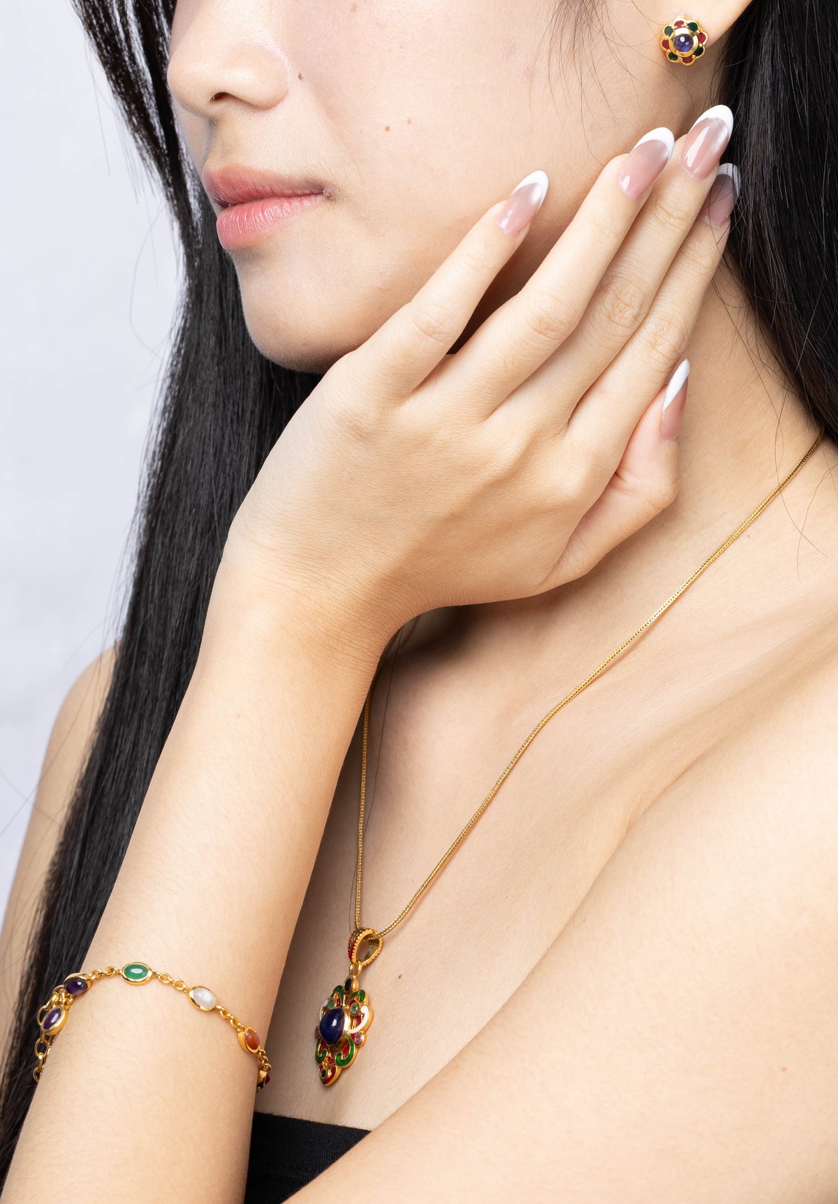 A woman with long black hair models a matching gold jewelry set, including a delicate necklace with a dark amber and green stone pendant, a gold bracelet, and a small gold stud earring, against a light gray background.