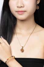 A close-up of a woman wearing a gold necklace with a round, colorful floral pendant, a matching gold bracelet, and a matching earring.