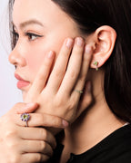 Close-up of a woman wearing a gold ring with a purple and yellow flower-shaped gemstone and a matching gold stud earring with a green stone.