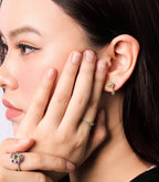 Close-up of a woman wearing a gold ring with a purple and yellow flower-shaped gemstone and a matching gold stud earring with a green stone.