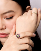 A close-up lifestyle shot of a woman resting her head on her hand, showcasing a silver ring with a purple and green gemstone cluster on her index finger, and another gold ring on her middle finger.