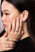 Close-up of a woman wearing delicate jewelry, including a small silver dangle earring, a silver ring with green stones, and a ring with a purple and gold floral design.