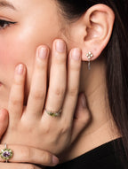 Close-up of a woman wearing delicate jewelry, including a small silver dangle earring, a silver ring with green stones, and a ring with a purple and gold floral design.