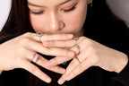 A close-up lifestyle shot of a woman wearing multiple delicate rings on her fingers, with her hands crossed in front of her face. 