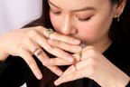 A close-up lifestyle shot of a woman wearing multiple rings on her fingers, including a prominent ring with a purple and green gemstone cluster, a silver band with purple stones, and a green stone accent ring.