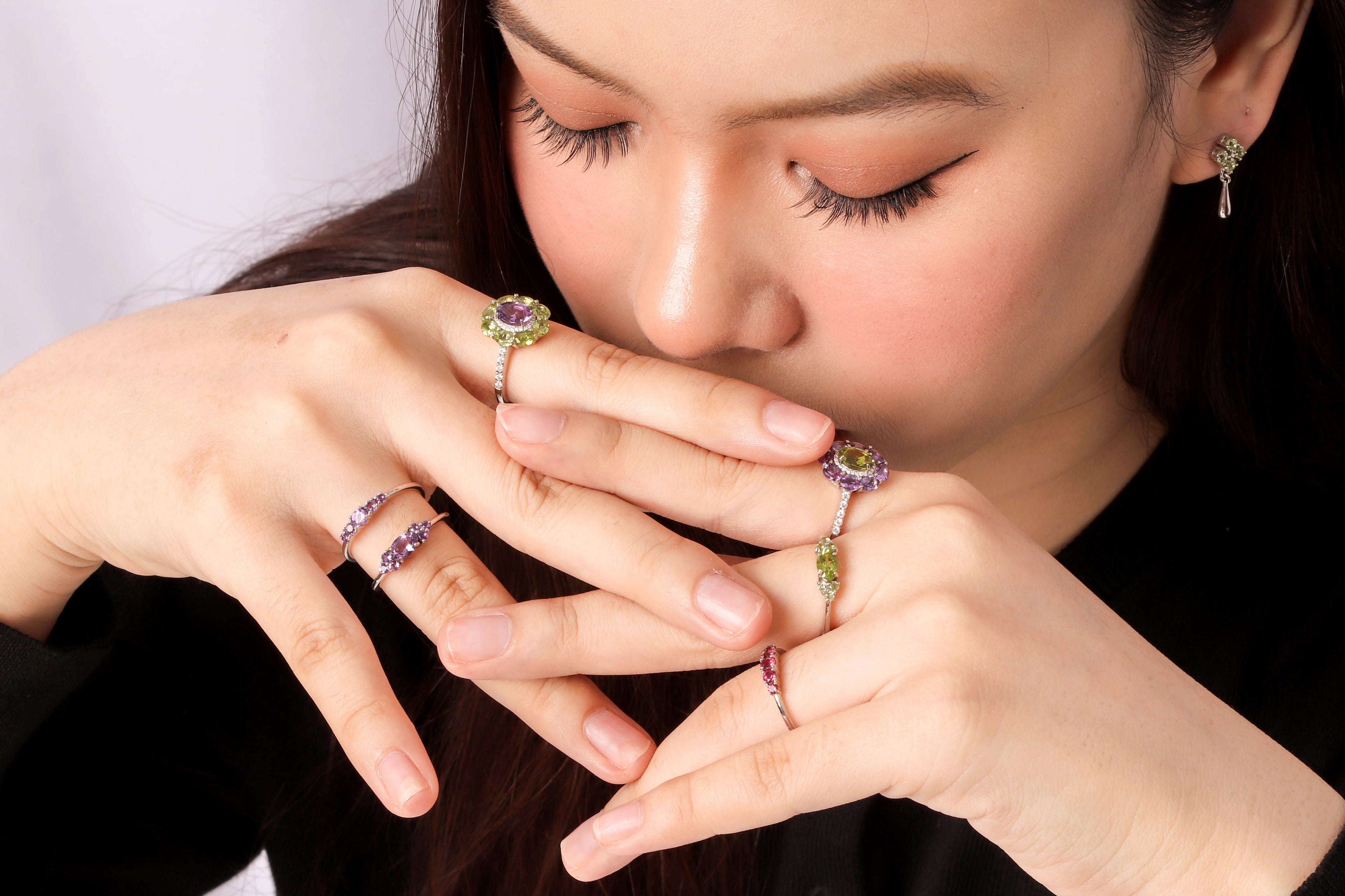 A close-up lifestyle shot of a woman wearing multiple rings on her fingers, including a prominent ring with a purple and green gemstone cluster, a silver band with purple stones, and a green stone accent ring.