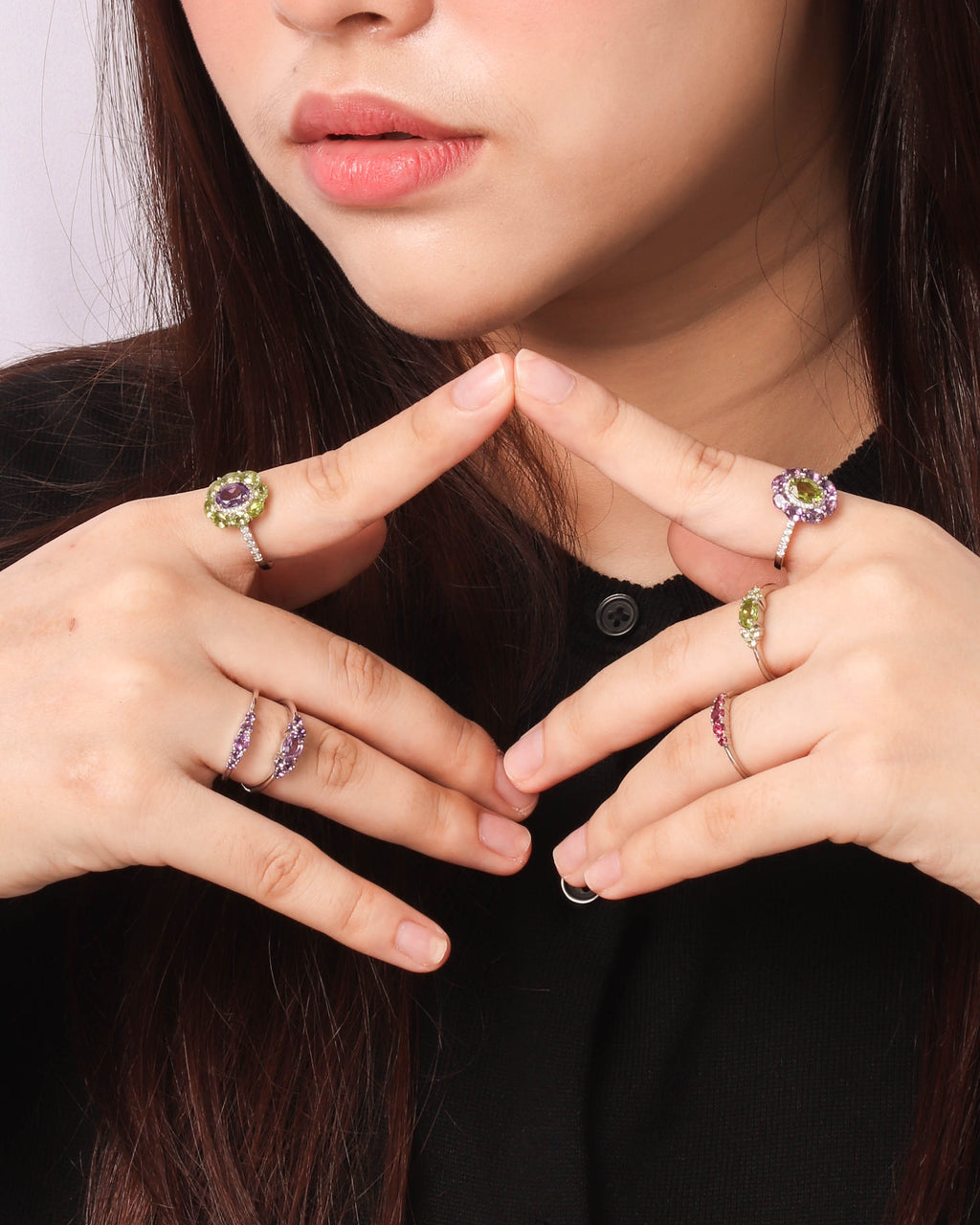 Close-up of a person wearing multiple silver rings with colorful gemstones, including purple and green accents, styled with a black top against a light background.