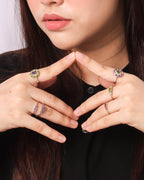 Close-up of a person wearing multiple silver rings with colorful gemstones, including purple and green accents, styled with a black top against a light background.
