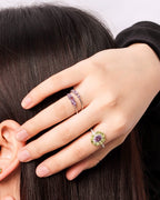 A close-up lifestyle shot of a woman with long dark hair wearing multiple colorful rings and a matching earring, with her hands touching her face and neck against a light gray background.