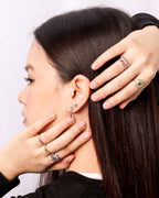 A close-up of a woman with long dark hair wearing multiple colorful gemstone rings and a small green stud earring, with her hands touching her hair and ear against a plain light background.