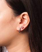 A close-up of a model's ear wearing a small huggie hoop earring with dark blue stones and a matching flower-shaped stud earring with a red flower center and green leaves.