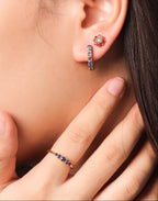 A close-up of a model's ear wearing a small floral stud earring and a vertical huggie hoop earring, with a matching ring on her finger.