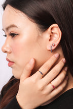 Close-up of a woman wearing a small hoop earring with pink stones and a blue stone, along with matching rings on her fingers, shown in profile against a light gray background.
