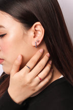 Close-up of a woman wearing small hoop earrings with pink gemstones and matching rings on her fingers, shown against a plain light background.