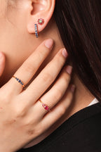 Close-up of a woman wearing small hoop earrings with pink gemstones and matching rings on her fingers, shown against a plain light background.