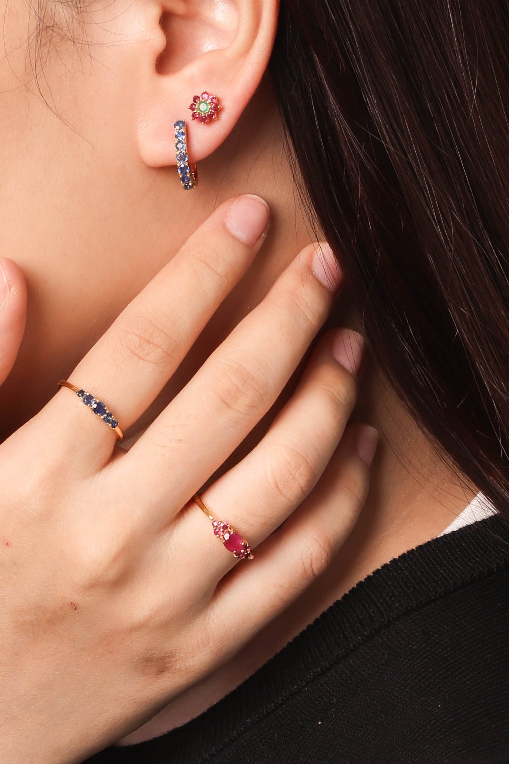 Close-up of a woman wearing small hoop earrings with pink gemstones and matching rings on her fingers, shown against a plain light background.