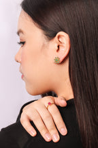 A close-up side profile of a woman with long dark hair, wearing a small green and gold floral stud earring and a matching gold ring with a red stone.