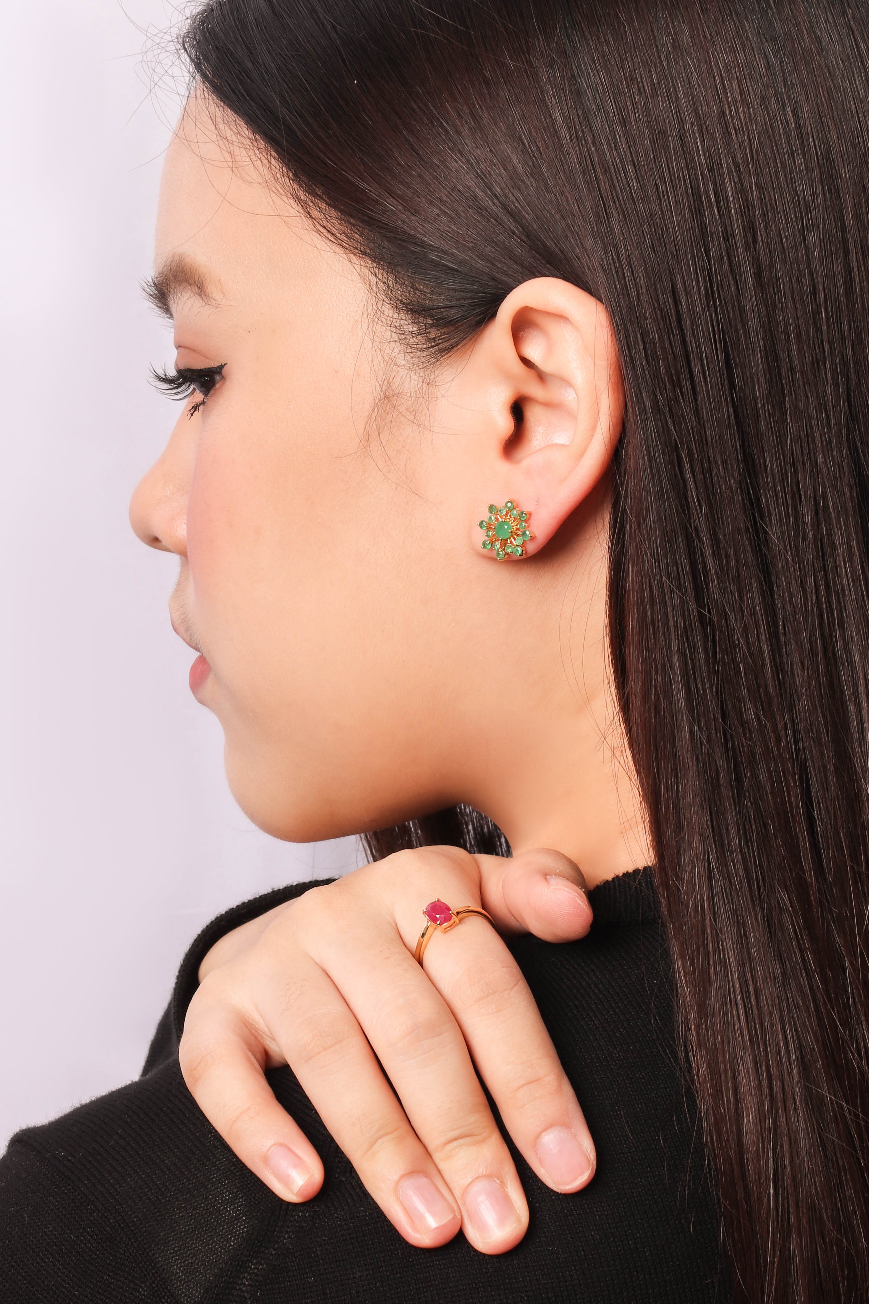 A close-up side profile of a woman with long dark hair, wearing a small green and gold floral stud earring and a matching gold ring with a red stone.