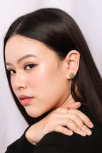 A close-up portrait of a woman with long dark hair, wearing a small green four-leaf clover stud earring and a gold ring with a red gemstone.