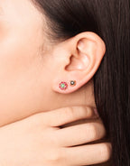 Close-up side profile of a woman wearing a gold ring with a green and red floral design on her finger and matching stud earrings on her earlobe.