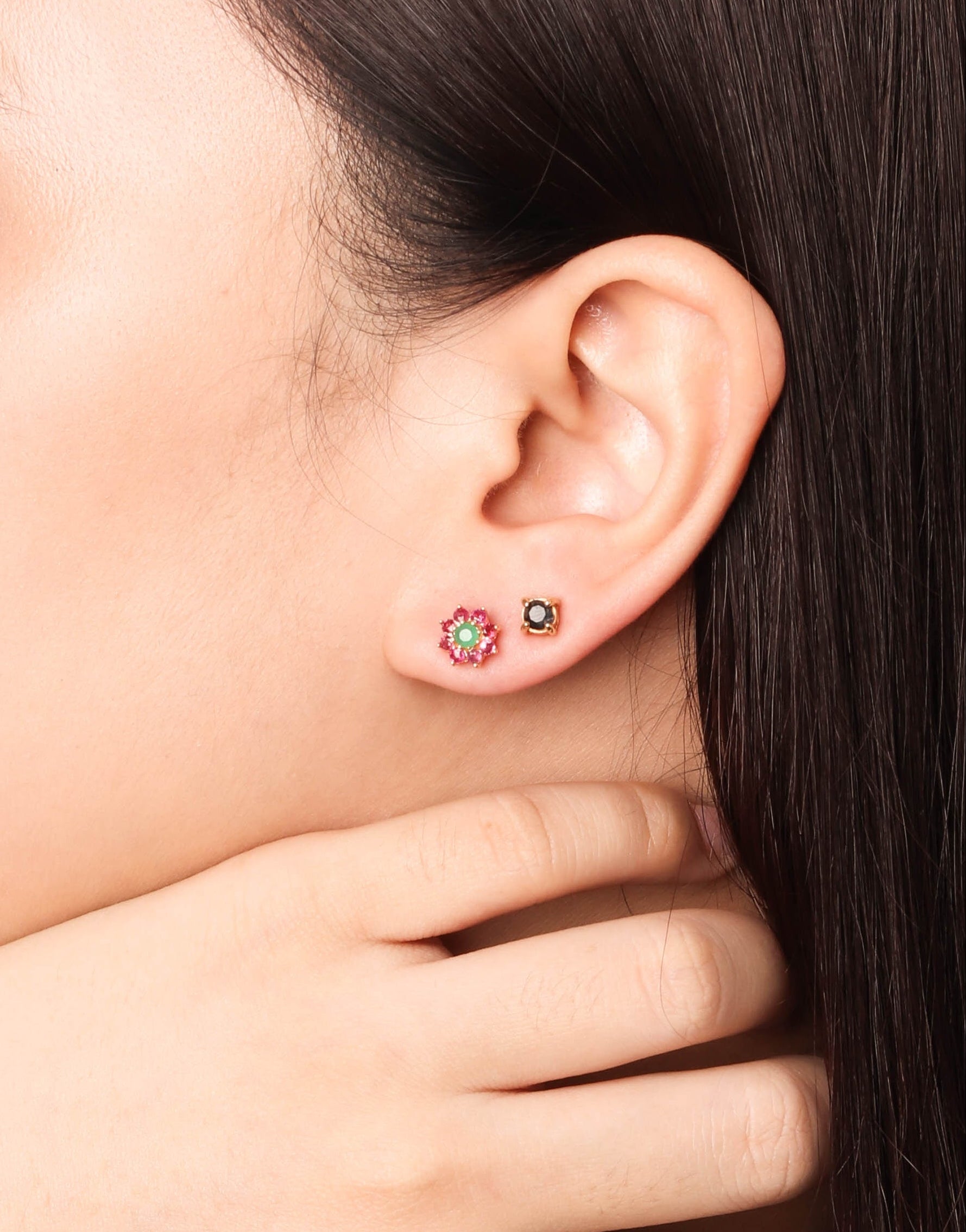 Close-up side profile of a woman wearing a gold ring with a green and red floral design on her finger and matching stud earrings on her earlobe.