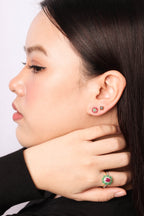 Close-up side profile of a woman wearing a gold ring with a green and red floral design on her finger and matching stud earrings on her earlobe.