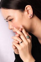 Close-up of a woman wearing small stud earrings and rings with green and red stones on silver settings, against a light gray background.