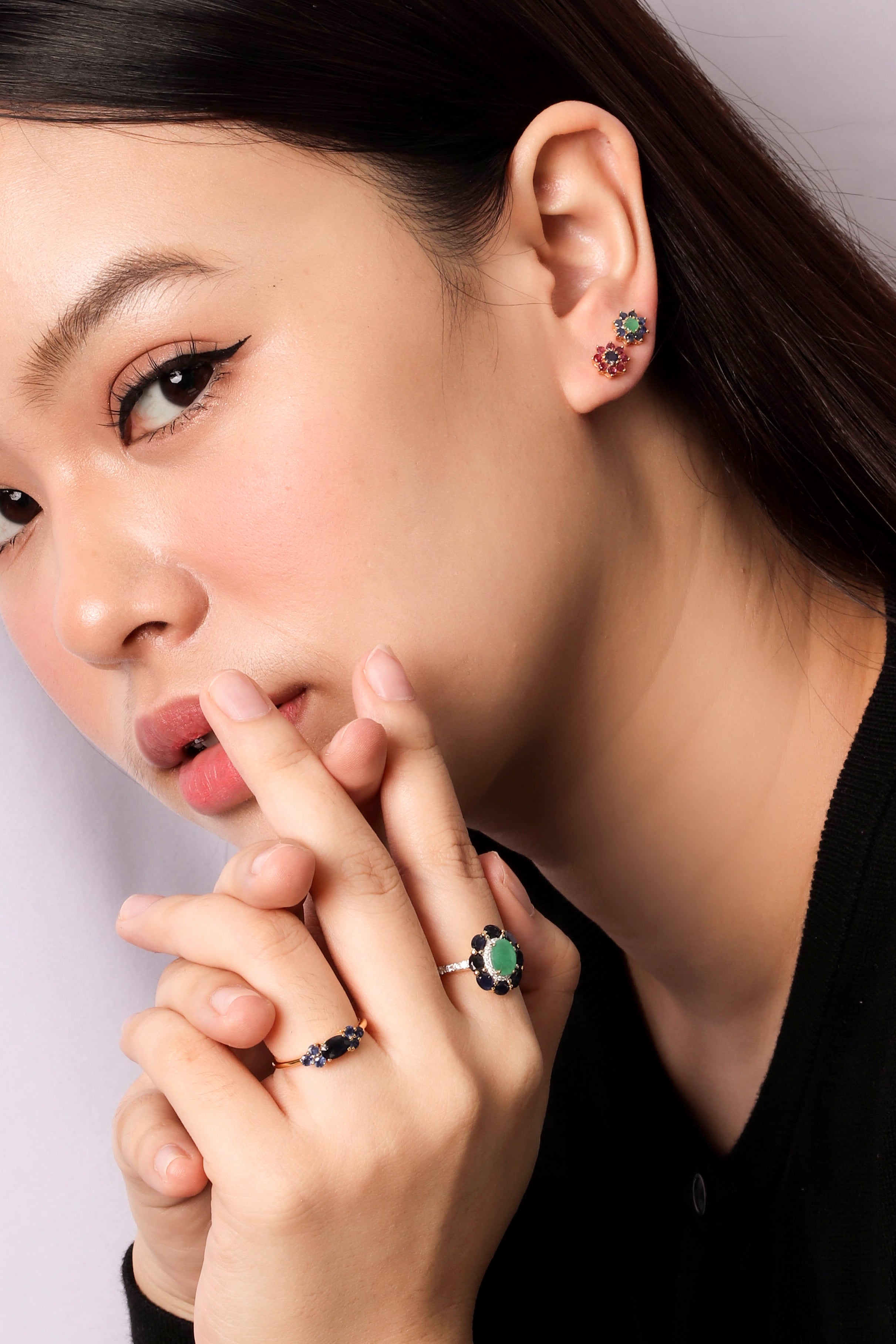 Close-up of a woman wearing a silver ring with a green flower-shaped stone and small black stones, along with matching stud earrings, against a light gray background.