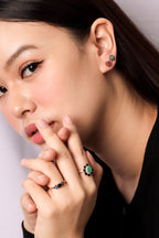 Close-up of a woman wearing a silver ring with a green flower-shaped stone and small black stones, along with matching stud earrings, against a light gray background.