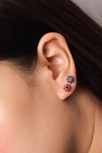Close-up of a person's ear wearing two small flower-shaped stud earrings, one with green stones and one with red stones.