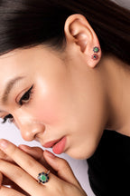 Close-up of a woman wearing a silver ring with a green and black floral design on her finger, along with matching stud earrings in her ear, against a light background.