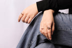 A close-up of a person's hands wearing multiple rings, including a prominent ring with a purple stone and a smaller gold band, while sitting in gray jeans.