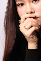 A close-up of a woman wearing gold rings with small clear gemstones on her fingers, set against a light gray background.