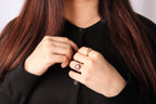 A close-up of a person wearing a black cardigan and jewelry, focusing on a gold ring with a floral cluster setting on their finger.