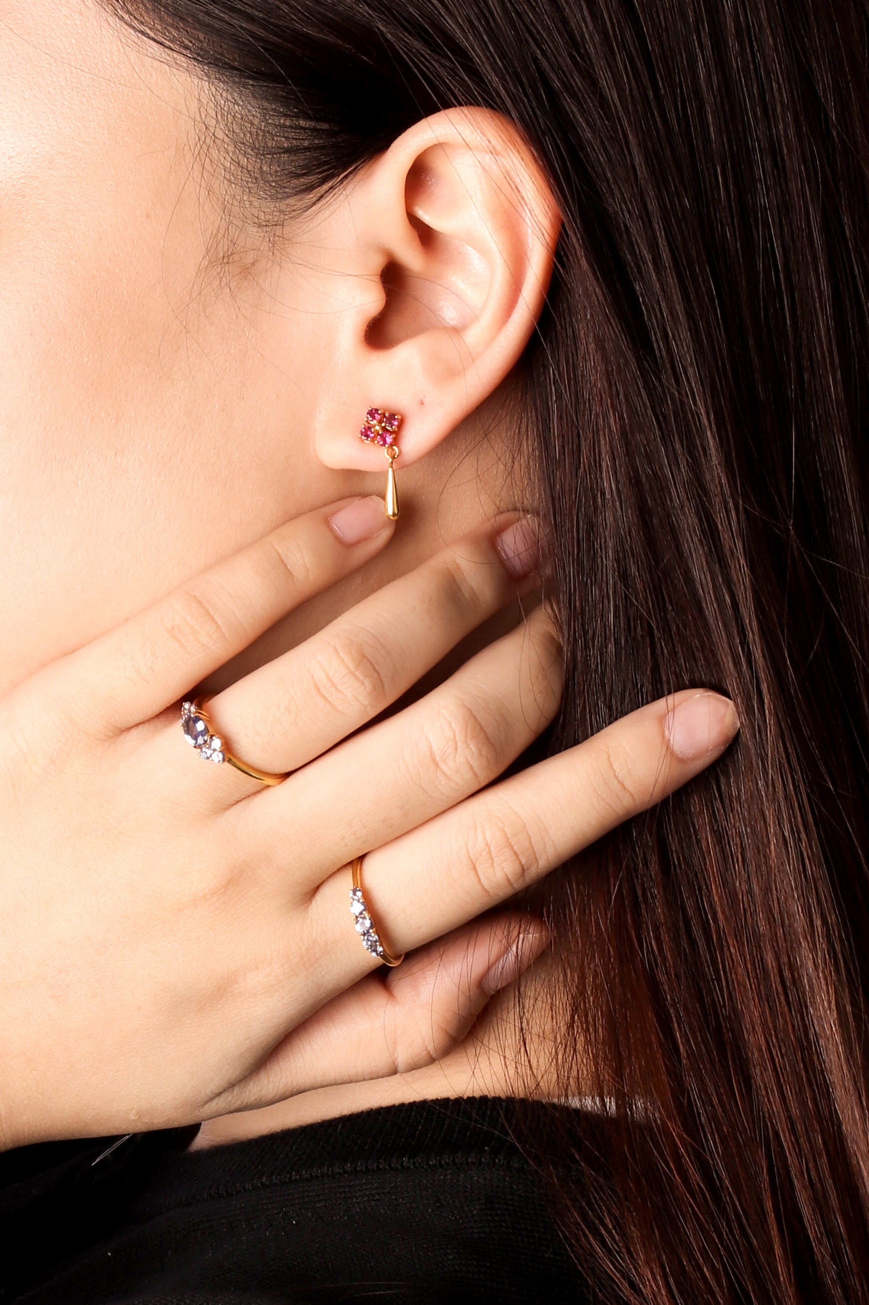 A close-up of a woman's ear and hand wearing a delicate gold huggie hoop earring with a small red gemstone at the top, along with two gold rings on her fingers.