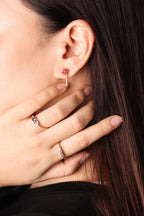 Close-up of a person's ear wearing a small gold hoop earring with a pink gemstone, with their hand resting near their face. 