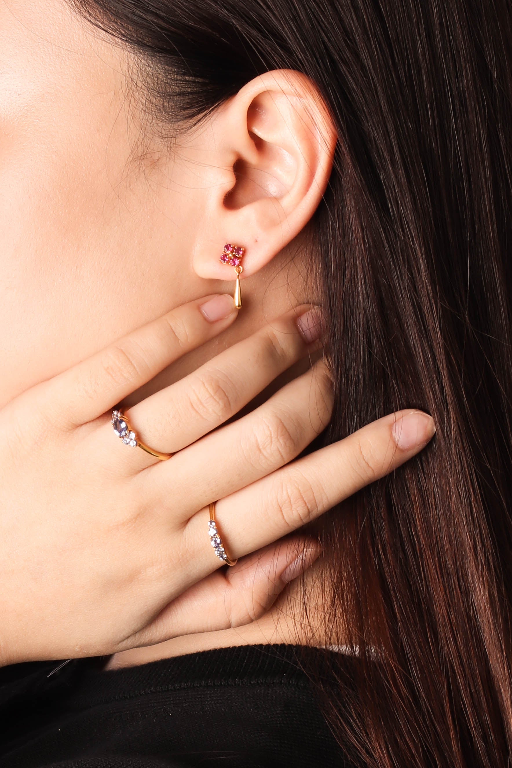 Close-up of a person's ear wearing a small gold hoop earring with a pink gemstone, with their hand resting near their face. 