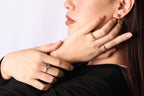 Close-up of a woman wearing multiple rings and an earring, with her hands resting on her neck against a plain light background.