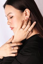 A close-up lifestyle shot of a woman with dark hair wearing several rings and an earring, including a prominent ring with a pinkish-purple heart-shaped gemstone on her finger.