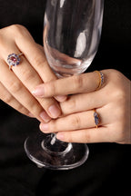 Close-up of two hands holding a clear champagne flute against a black background, wearing several rings including a prominent ring with a round red gemstone surrounded by small clear stones.