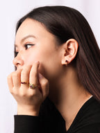 A close-up side profile of a woman with long dark hair, wearing small gold hoop earrings and two delicate gold rings, one of which features a small pink gemstone. She is wearing a black top against a clean white background.