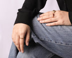 Close-up of a person's hands resting on their lap, wearing two delicate gold rings with small colorful gemstone accents. One ring features a tiny red stone, and the other has a small yellow/green stone.