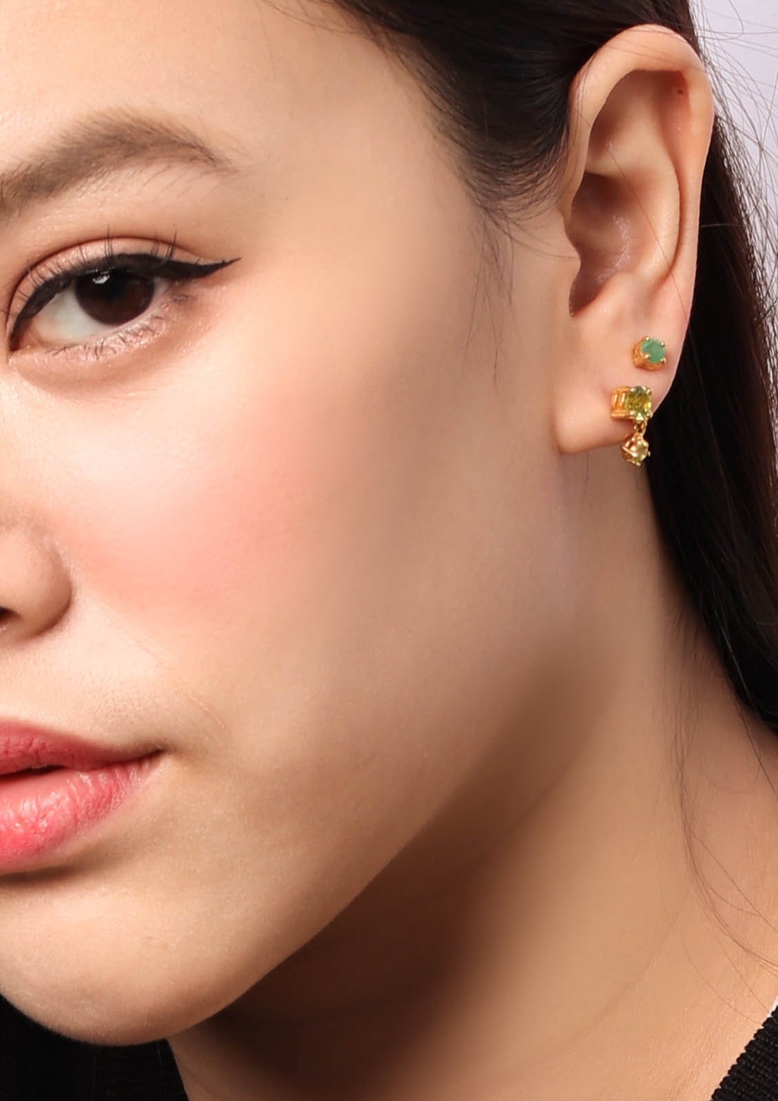 Close-up of a woman's ear wearing a pair of small gold stud earrings with green and yellow-green stones.